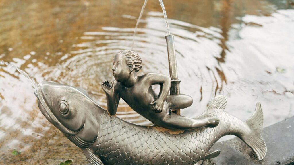 Artistic metal sculpture of child riding fish in an outdoor fountain in Zurich park.