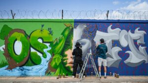 Two artists working on a vibrant mural on an urban wall. Captured in daylight showing detailed graffiti and artistic expression.