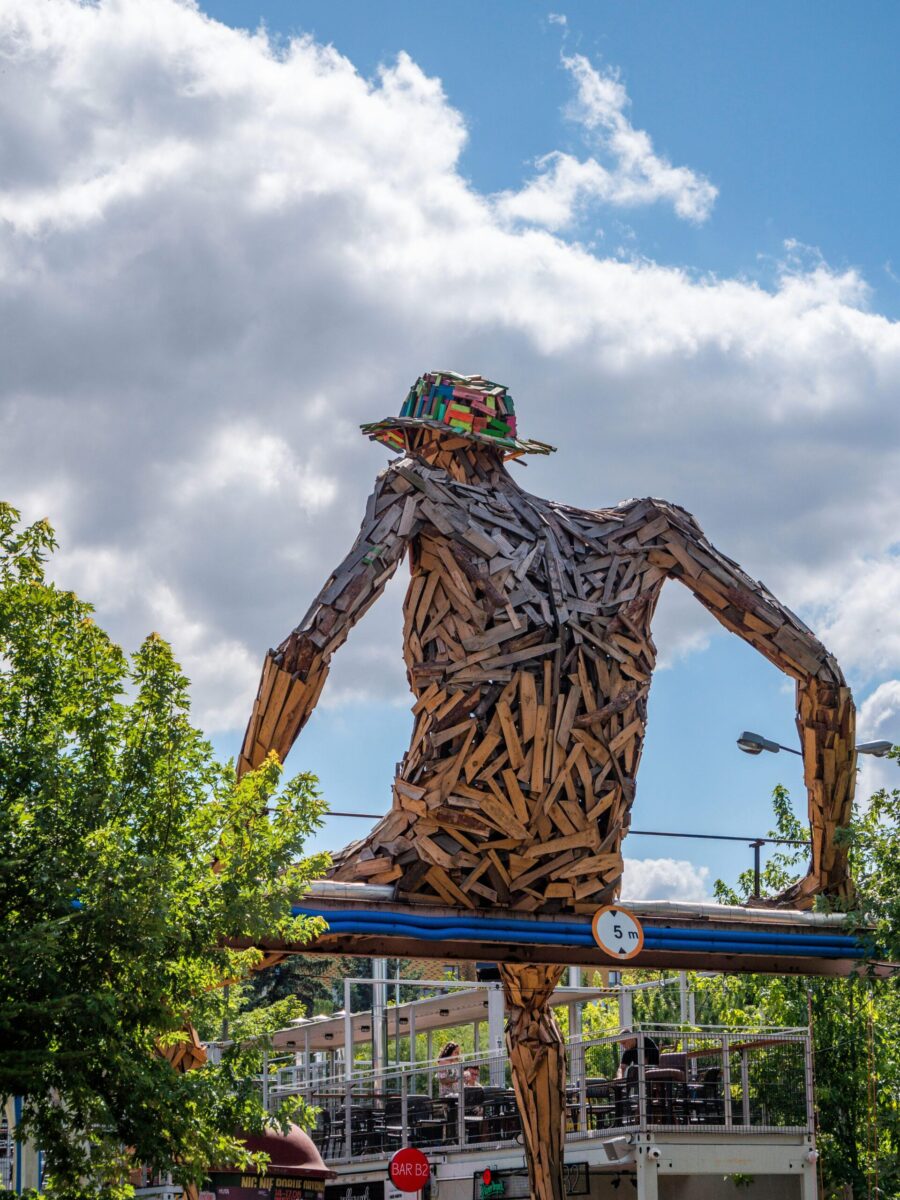 Artistic sculpture made of wood shaped into a human figure wearing a colorful hat, set against a cloudy sky.